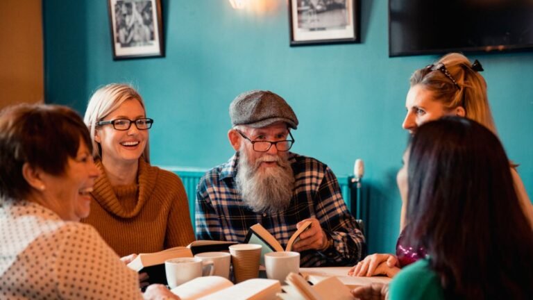 Diverse group of adults engaged in animated book club discussion around cafe table with notebooks and coffee